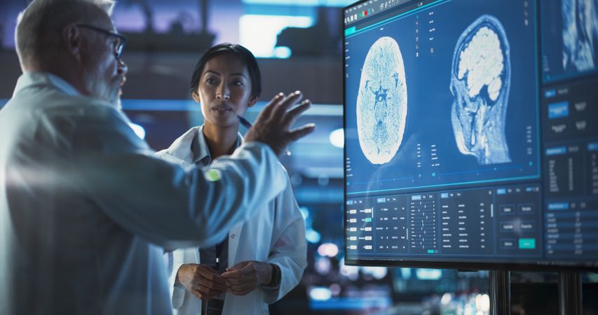 Neuroscience Laboratory Meeting: Female Asian Scientist And Male Surgeon Discussing MRI Brain Scan On TV Display.