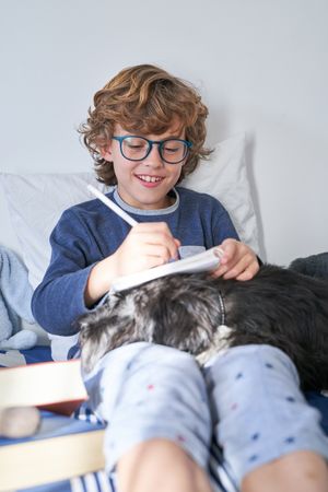 Cheerful boy doing homework on bed with dog