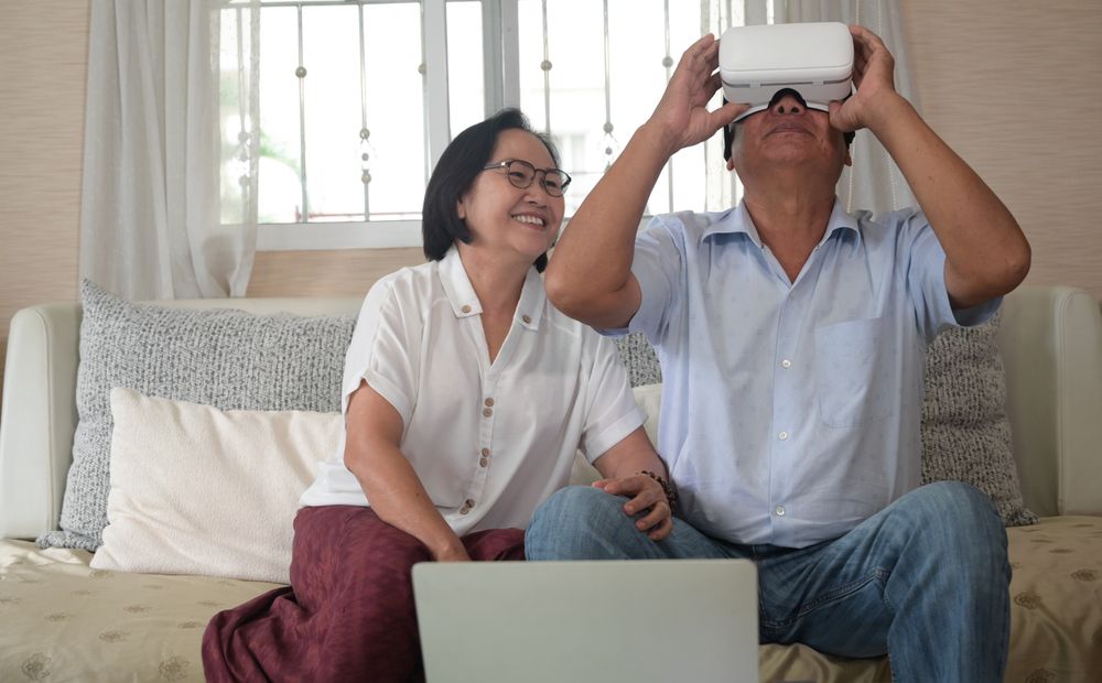 An older man wearing a VR headset and his wife sitting side by side having a video call on the sofa in the house.