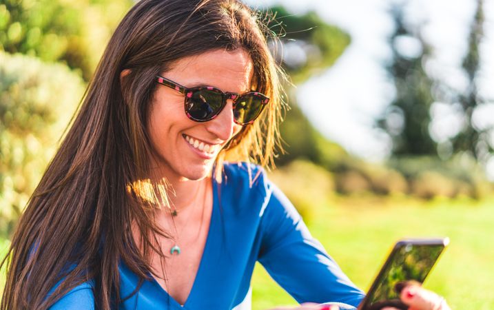 portrait of young woman with sunglasses using smartphone sitting on grass in park