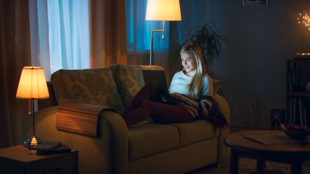 In the Evening Young Woman Lies on the Couch with a Laptop on Her Knees Typing