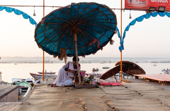 Indian man in light textile sitting under umbrella near Ganga river in Uttar Pradesh, India