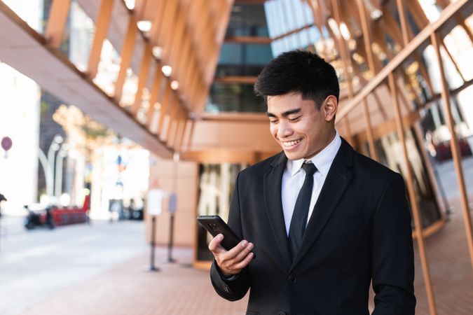 Cheerful Asian male entrepreneur browsing smartphone in city