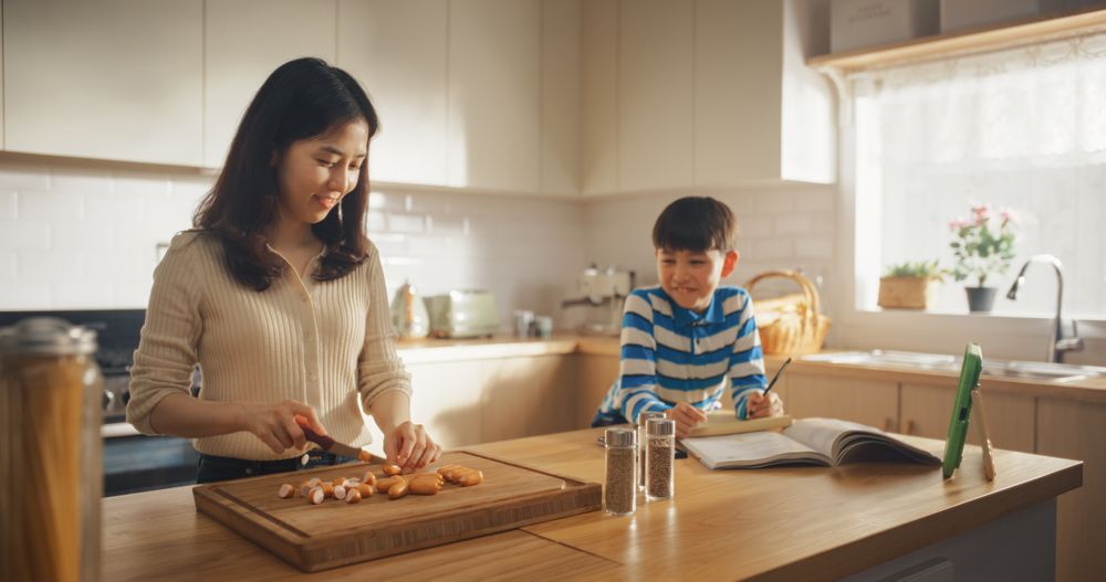 Portrait of a Korean Woman and her Son in the Kitchen at Home: Mother Preparing for Cooking Lunch While Talking to her Cute Kid Who is Doing Homework Next to Her.