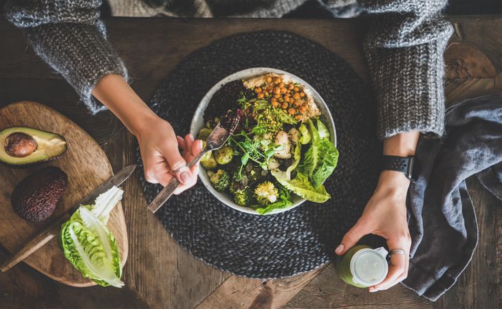 Healthy vegetarian bowl pictured on table with smoothie on side and hands with fork