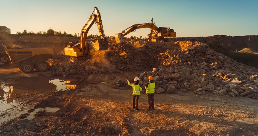 Aerial Drone Shot Of Construction Site On Sunny Evening: Industrial Excavators Digging Rocks And Loading Into A Truck. Engineer And Inspector Observing Process, Discussing New Real Estate Project