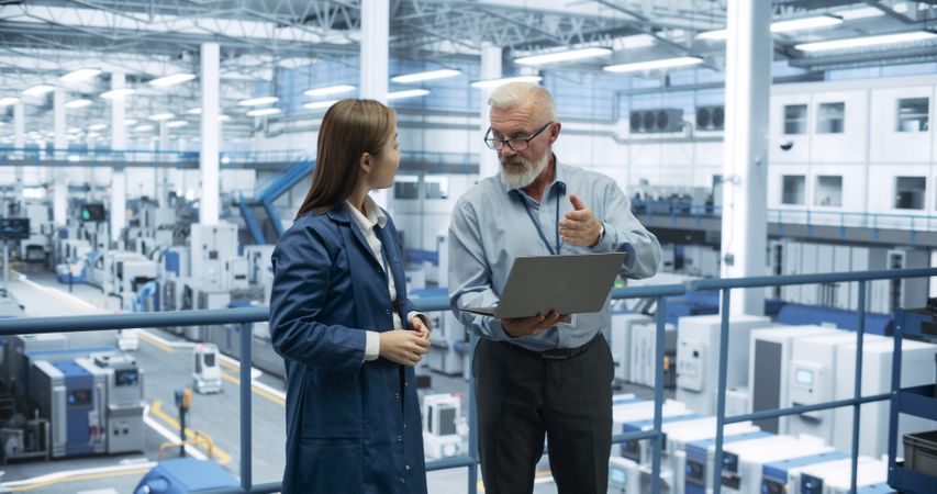 Two Diverse Engineers Standing on a Platform, Using Laptop Computer at an Electronics Factory. Machines are Producing Electronic Chips, Specialists Monitoring the Progress on Online Software