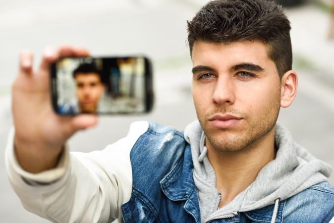 Serious man in jean jacket taking selfie outside