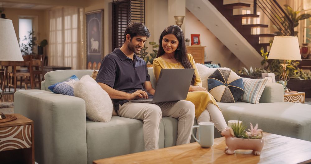 Portrait of Loving Young Indian Couple Enjoys Browsing Online Shopping Websites Together On A Laptop, While Sitting Cozily On Their Living Room Sofa.