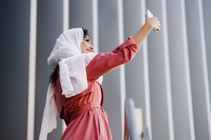 Female in headscarf and pink trench coat taking photo with her phone
