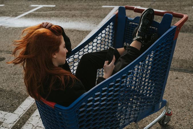 Smiling woman with smartphone in shopping trolley in parking lot