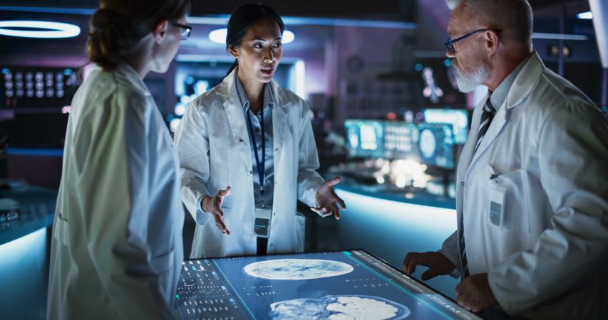 Modern Hospital Medical Research Center: Diverse Colleagues Gathered Around Interactive Touch Screen Table With MRI Scans Of Brain On Display.