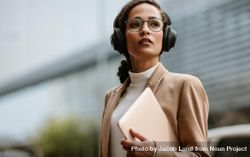 Businesswoman wearing headphones walking down the city street holding a laptop 5qyd15