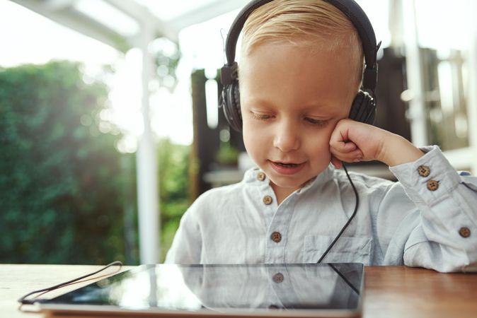 Blond boy using headphones and watching show or playing game on digital tablet