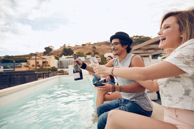 Outdoor shot of happy young people sitting on the edge of the pool drinking champagne