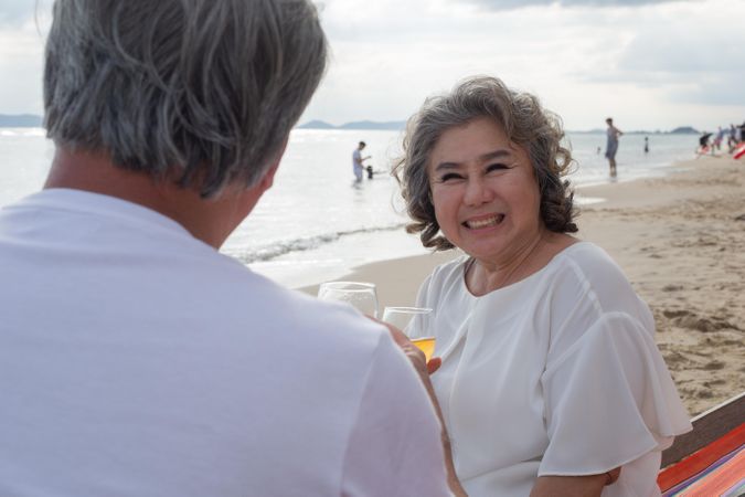Smiling Asian couple relaxing on the beach and toasting with wine