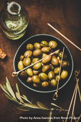 Bowl of olives on wooden table with garnish and bottle of olive oil, and skewers for serving 5rPNPb