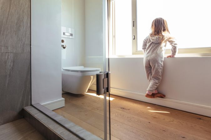 Little girl in bathroom standing by a big window and looking outside