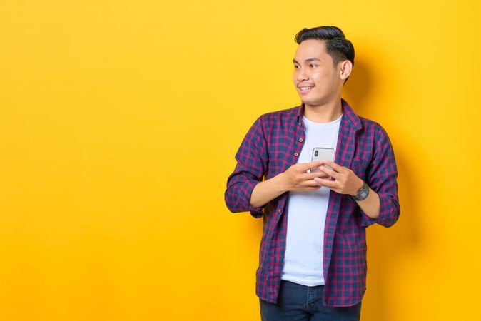 Smiling Asian in plaid shirt  holding mobile phone while looking away