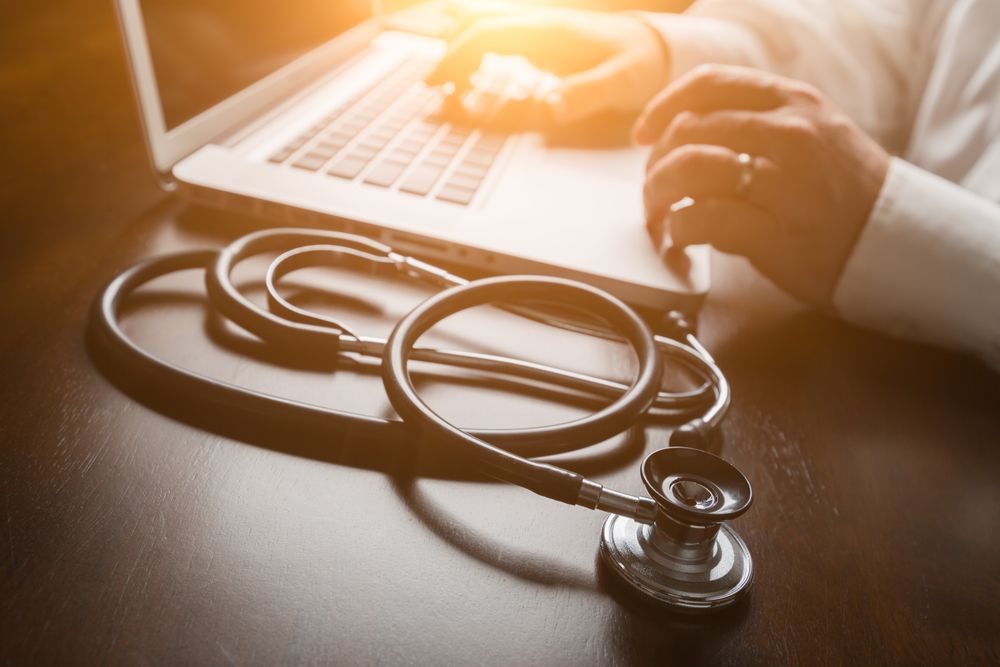 Medical Stethoscope Resting on Desk As Male Hands Type on Computer Keyboard.