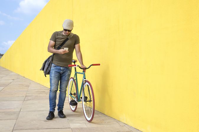 Man in hat and sunglasses walking along yellow wall and checking phone with bike