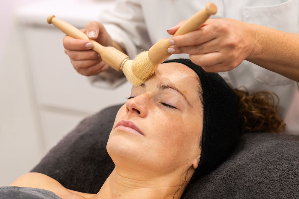 Crop professional cosmetologist cleaning face of female client with brushes in beauty salon