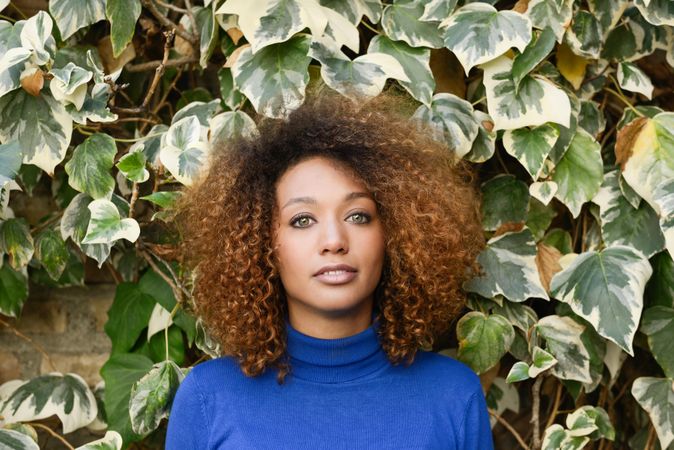 Woman with curly hair looking at camera in front of wall of leaves