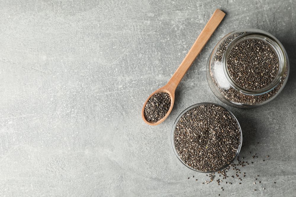 Glass bowl, jar and wood spoon with chia on grey background, copy space