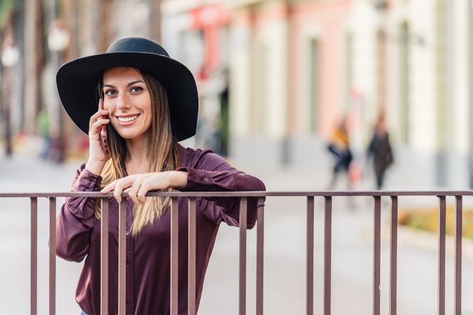 Charming woman in trendy outfit talking on smartphone and smiling at city street