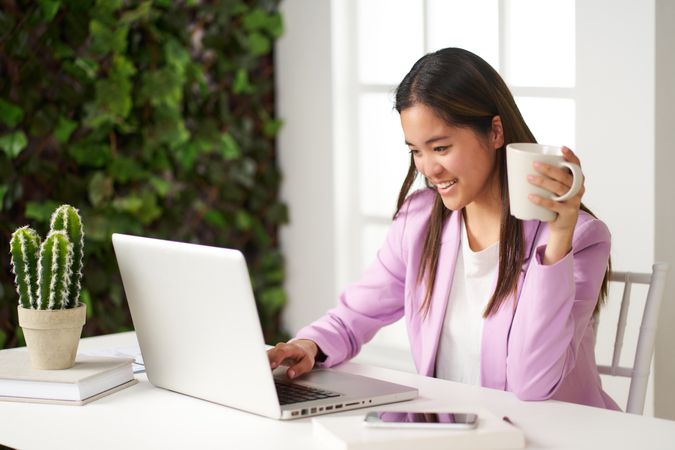 a young asian woman smiling working at the computer in the coworking space having coffee