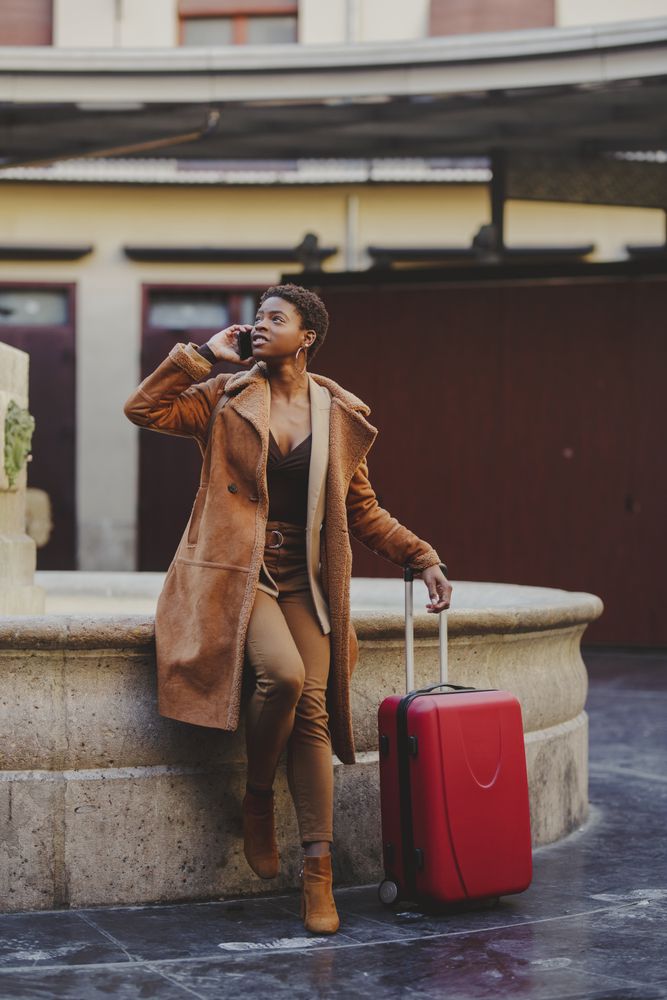 Stylish Black woman talking on smartphone with luggage on street