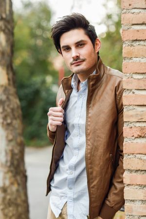 Man in leather coat leaning on brick wall outside