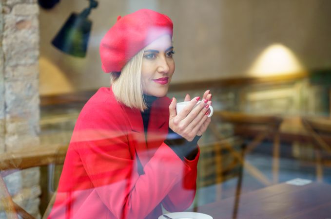 Through glass side view of attractive female in red elegant coat and beret sitting at table with cup of fresh coffee in light cafe