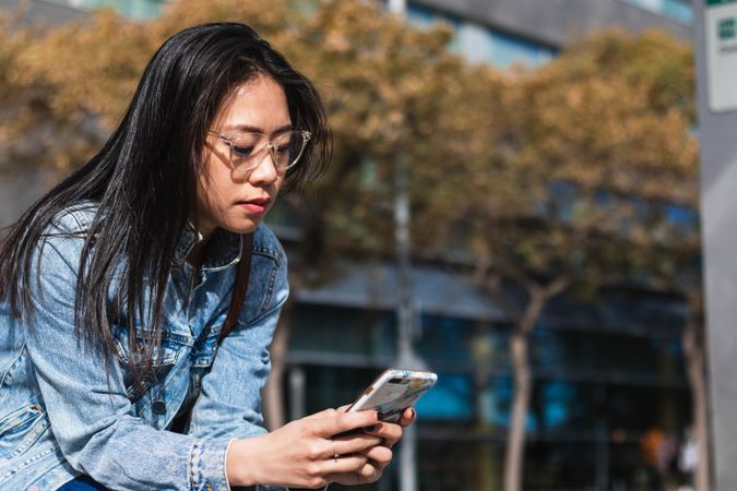 Long hair brunette Asian woman using mobile phone while sitting on a bench in the street