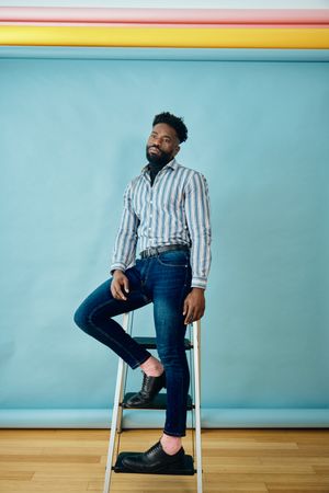 Serious Black male posing in blue studio in jeans and striped shirt on step ladder