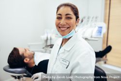 Happy dentist smiling at camera with male patient in the background bGlJA0
