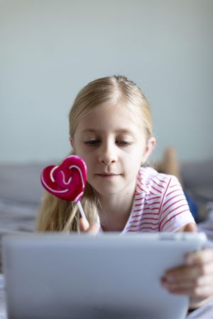 Young girl holding a pink heart shaped lollipop using a tablet
