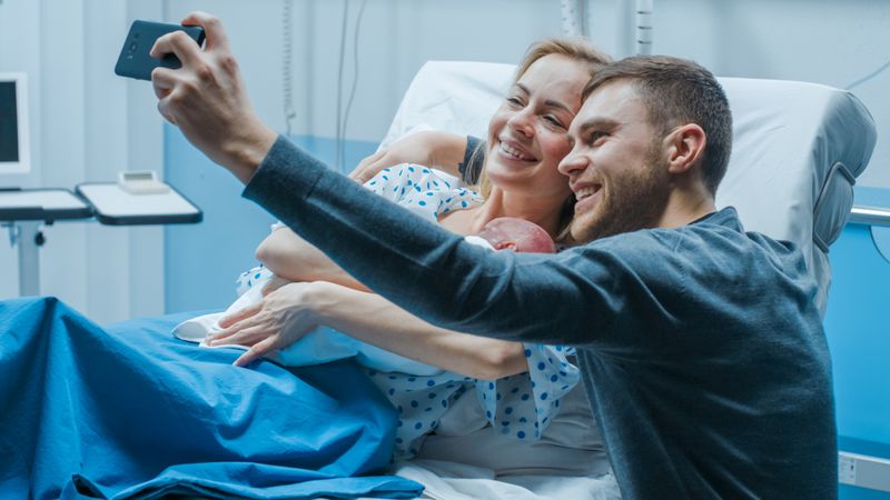 Father Takes Selfie of Him with His Wife Holding Newborn Baby while Lying on the Hospital Bed