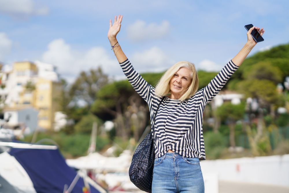 Older woman in jeans and striped shirt outside on sunny day with arms up in excitement