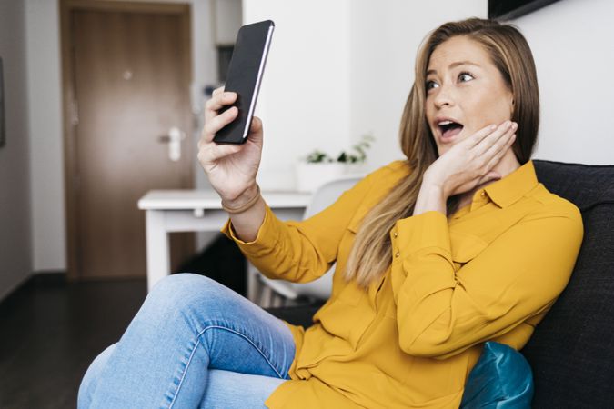 Surprised adult female sitting on sofa while using smartphone