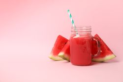 Glass jar of watermelon juice and slices on pink background