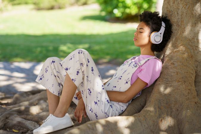 Woman in floral coveralls relaxing on roots of large tree in park listening to music