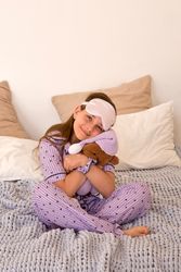 Girl hugging teddy bear on bed, wearing sleep mask.