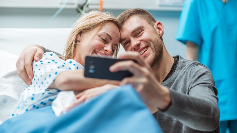Father Takes Selfie of Him with His Wife Holding Newborn Baby while Lying on the Hospital Bed