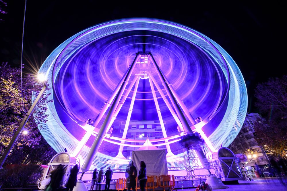 Slow shutter shot of purple lit ferris wheel at night with people queuing in front