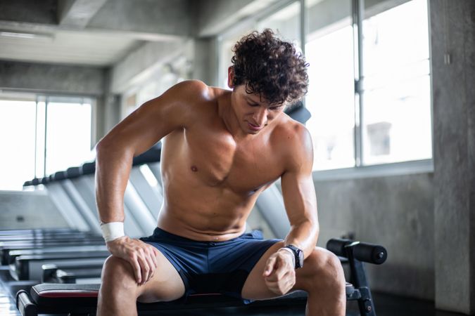 Muscular male sitting on workout bench in gym between sets