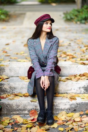 Female in warm winter clothes sitting among scattered fall leaves on park steps looking away