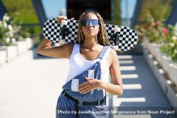 Female in denim overalls with checkered skateboard behind her head with one hand on stomach 4dxEl4