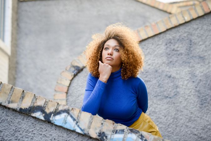 Thoughtful female with curly hair wearing bright blue shirt standing on outdoor stairs