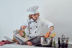 Positive boy sitting on counter and putting vegetables into sauc